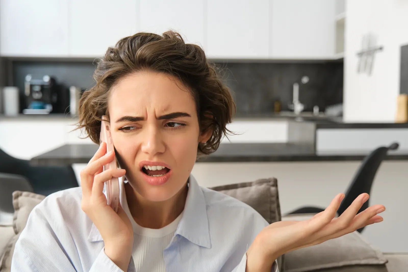 Frustrated woman on a phone call in her kitchen, shrugging with a confused expression