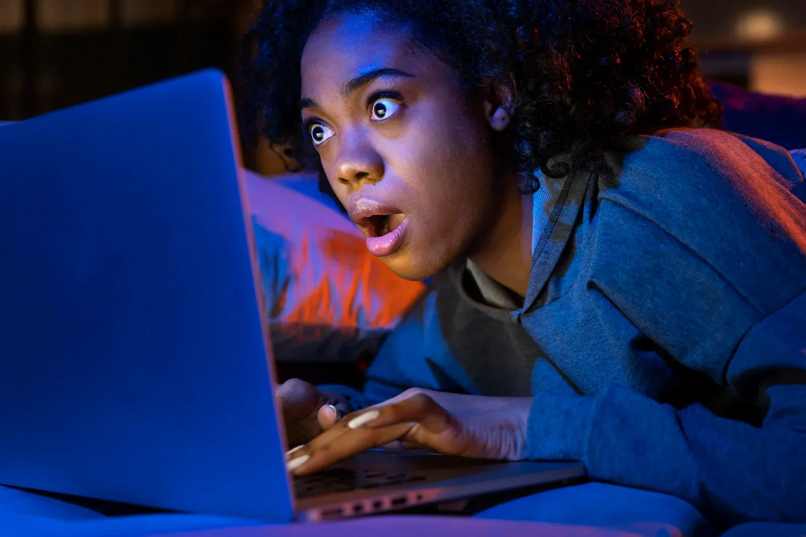 Young woman lying down looking shocked at a laptop screen lit blue in a dark room