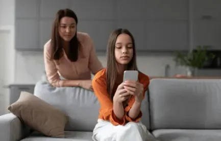 Mother watching over her daughter as the girl uses a smartphone on the couch
