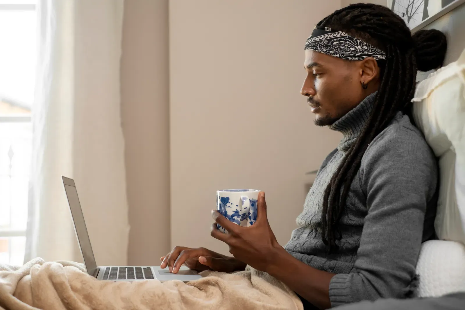 Man holding a patterned mug and typing on a laptop while relaxing in bed