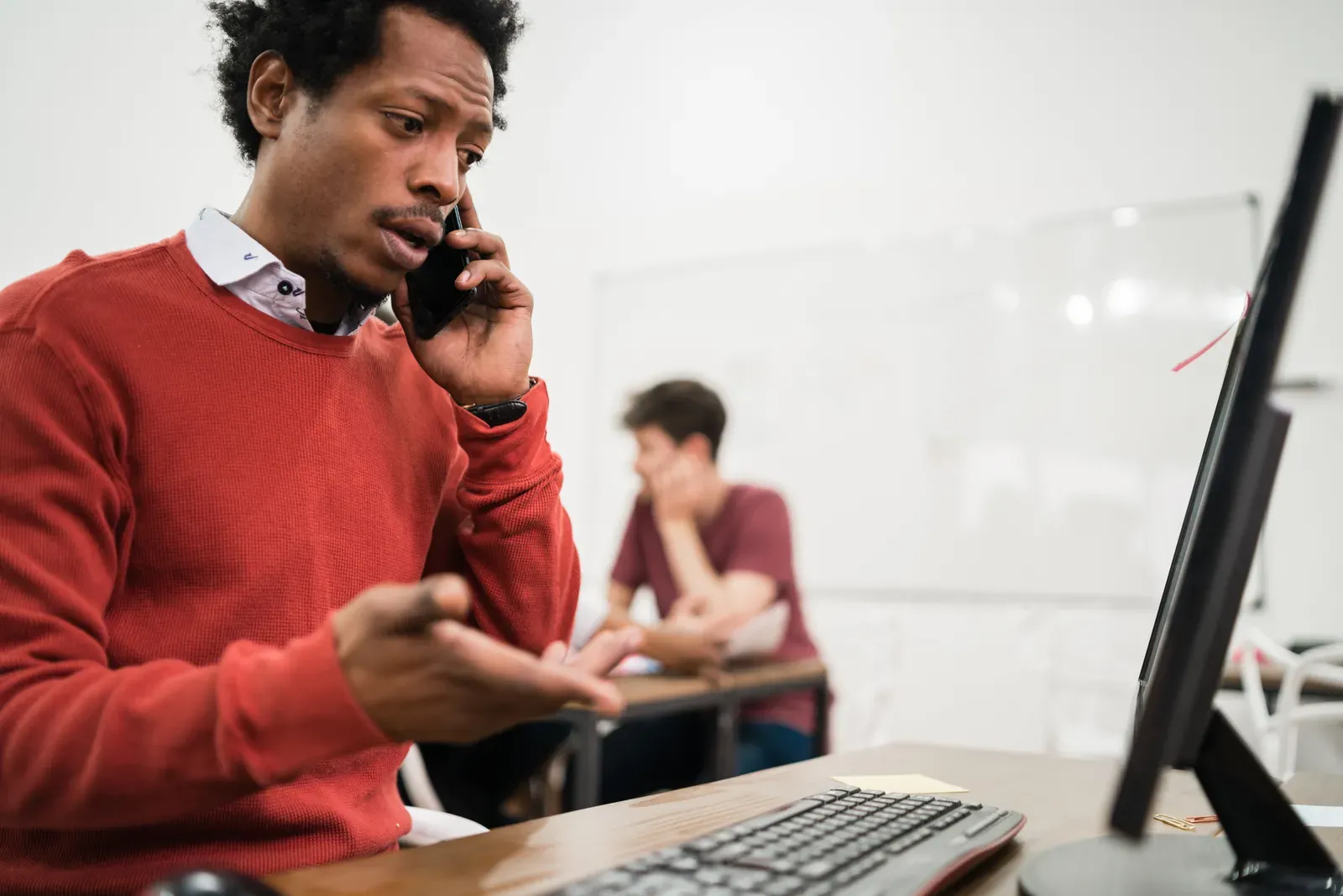 Frustrated man on the phone gesturing at a desktop monitor in an office