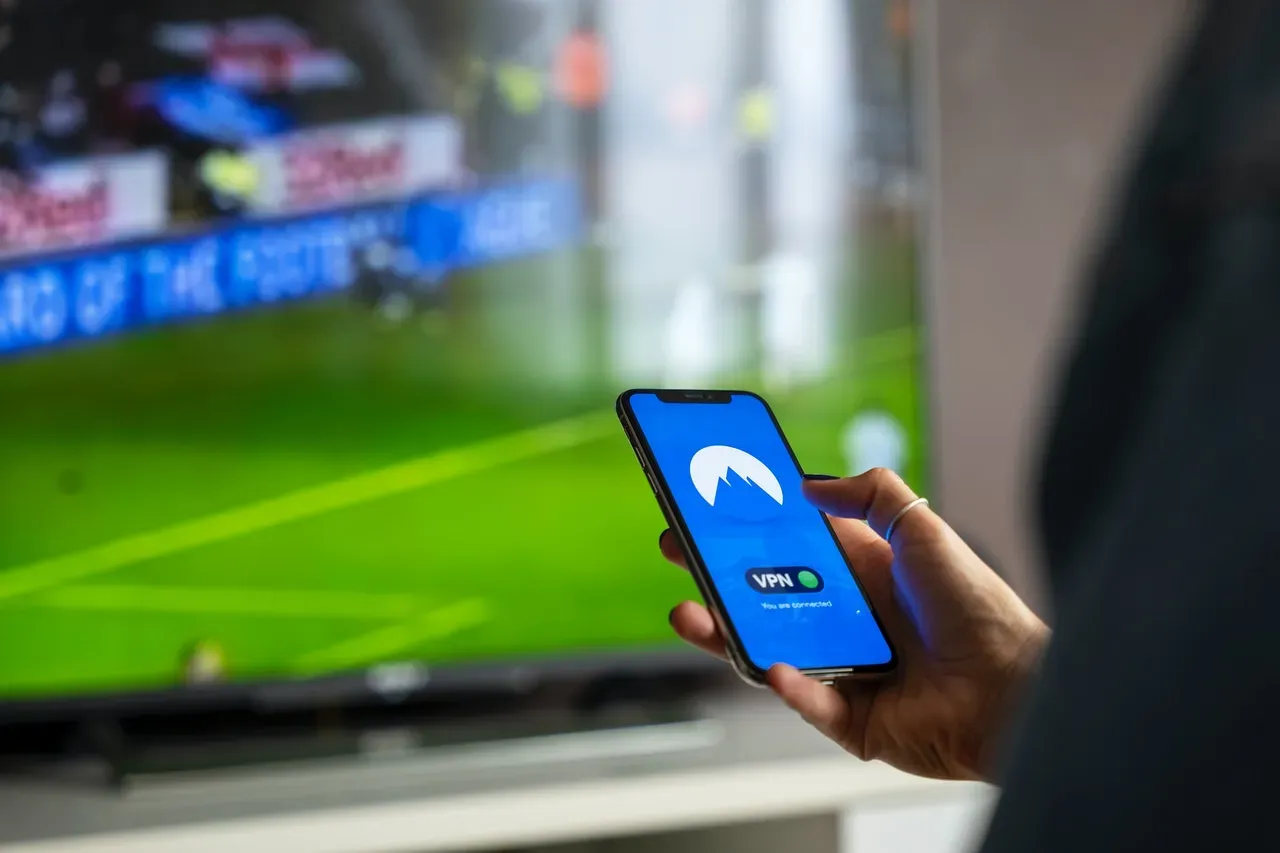 Hand holding a phone running a VPN app in front of a TV showing a soccer match