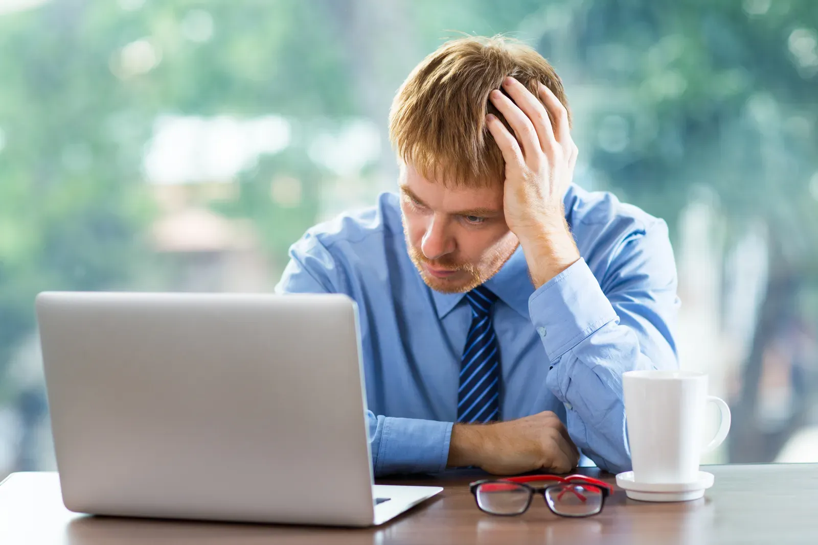 Stressed man in a shirt and tie holding his head while looking at a laptop at his desk